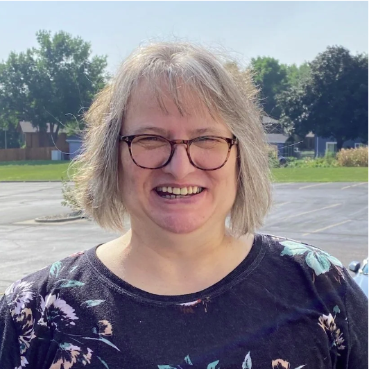 A woman with shoulder-length gray hair, glasses, and a floral shirt, smiling outdoors on a sunny day in an empty parking lot with trees in the background.