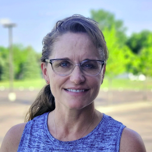 Woman with glasses, long brown hair, wearing a blue sleeveless top, smiling outdoors with greenery in the background.