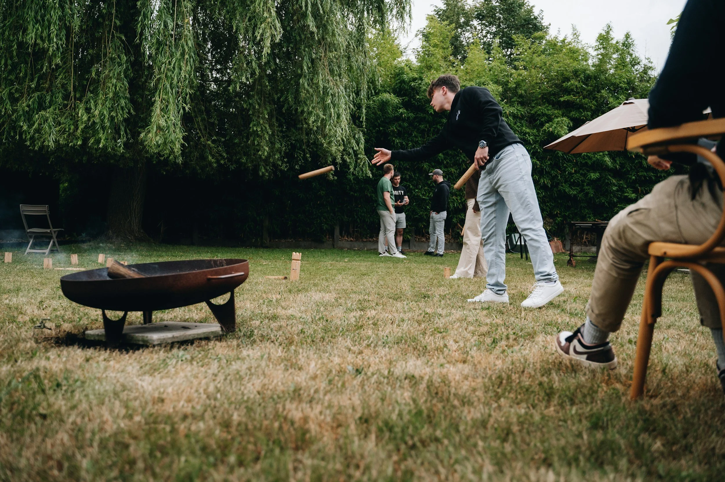 A group of people playing cornhole outdoors on a grassy lawn with trees in the background. One person is throwing a bean bag towards the cornhole board while others watch.