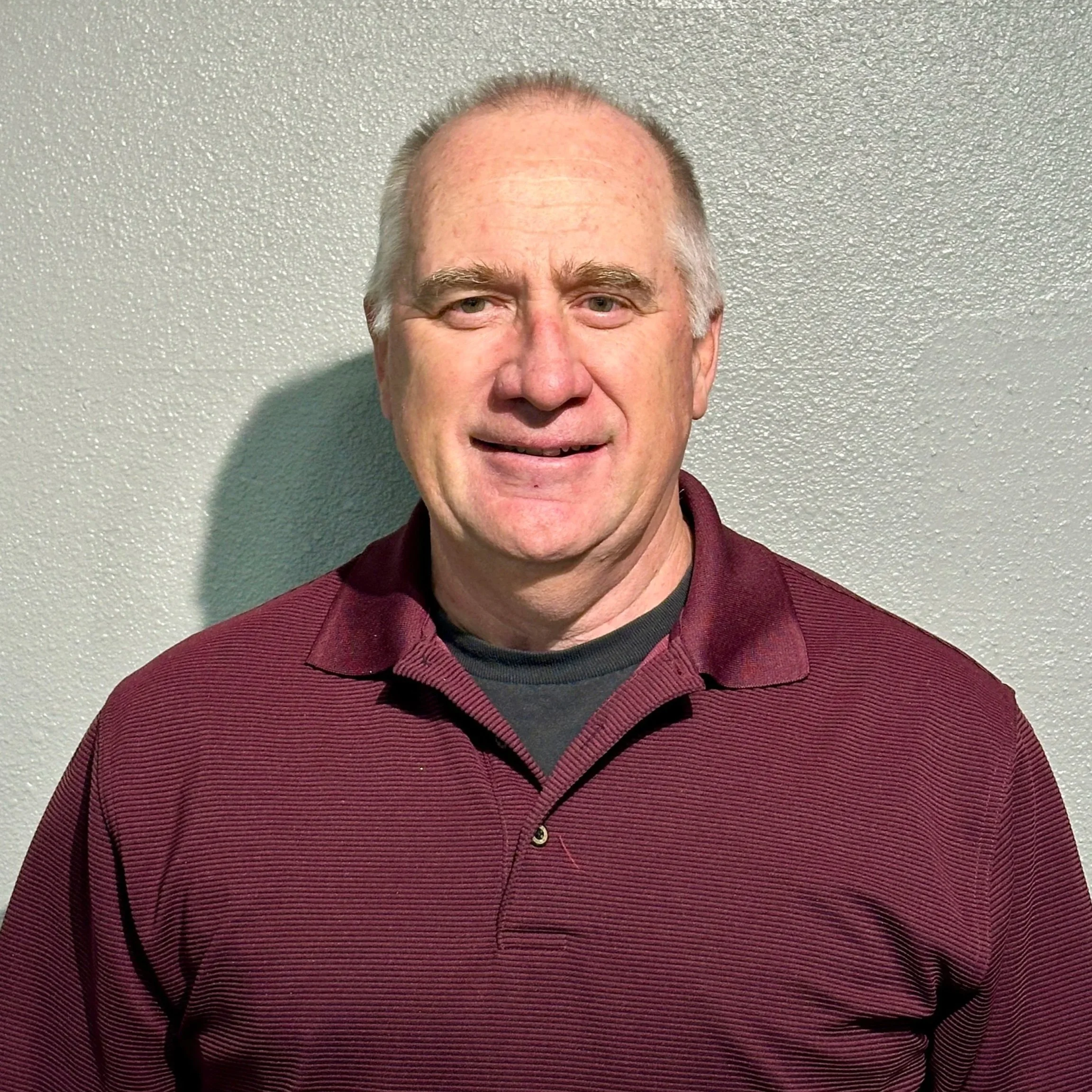 A middle-aged man with thinning light-colored hair, wearing a dark maroon collared shirt over a gray shirt, standing against a light textured wall.