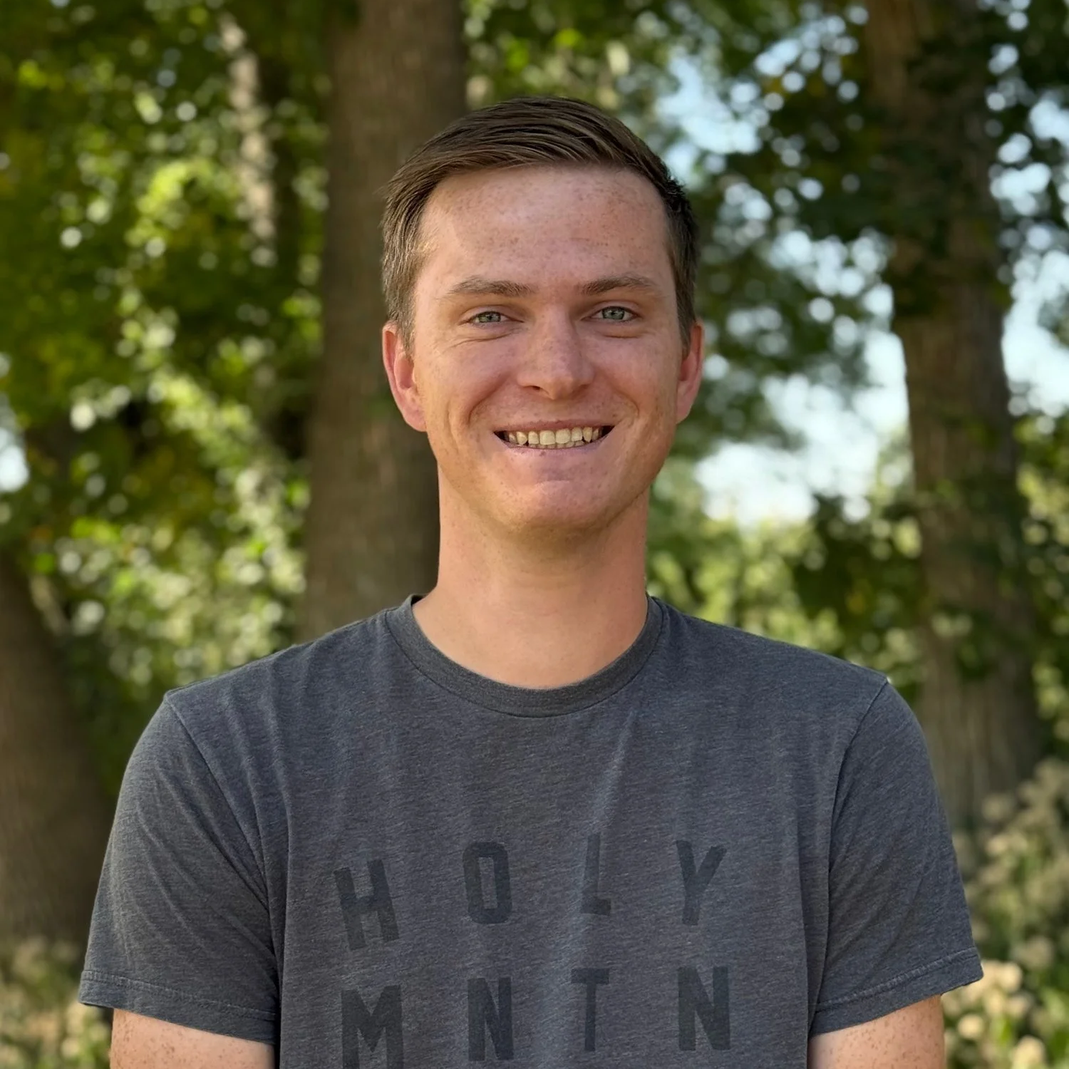 Young man with short brown hair, smiling outdoors with trees in the background, wearing a dark gray T-shirt.