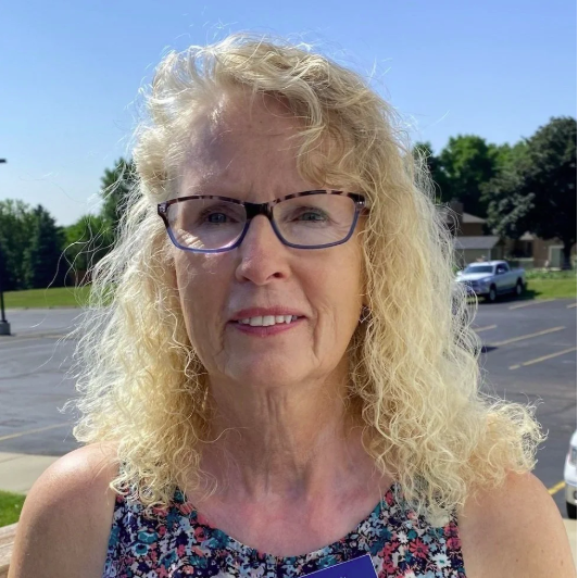 A woman with blonde curly hair and glasses smiling outdoors on a sunny day, with a parking lot and trees in the background.