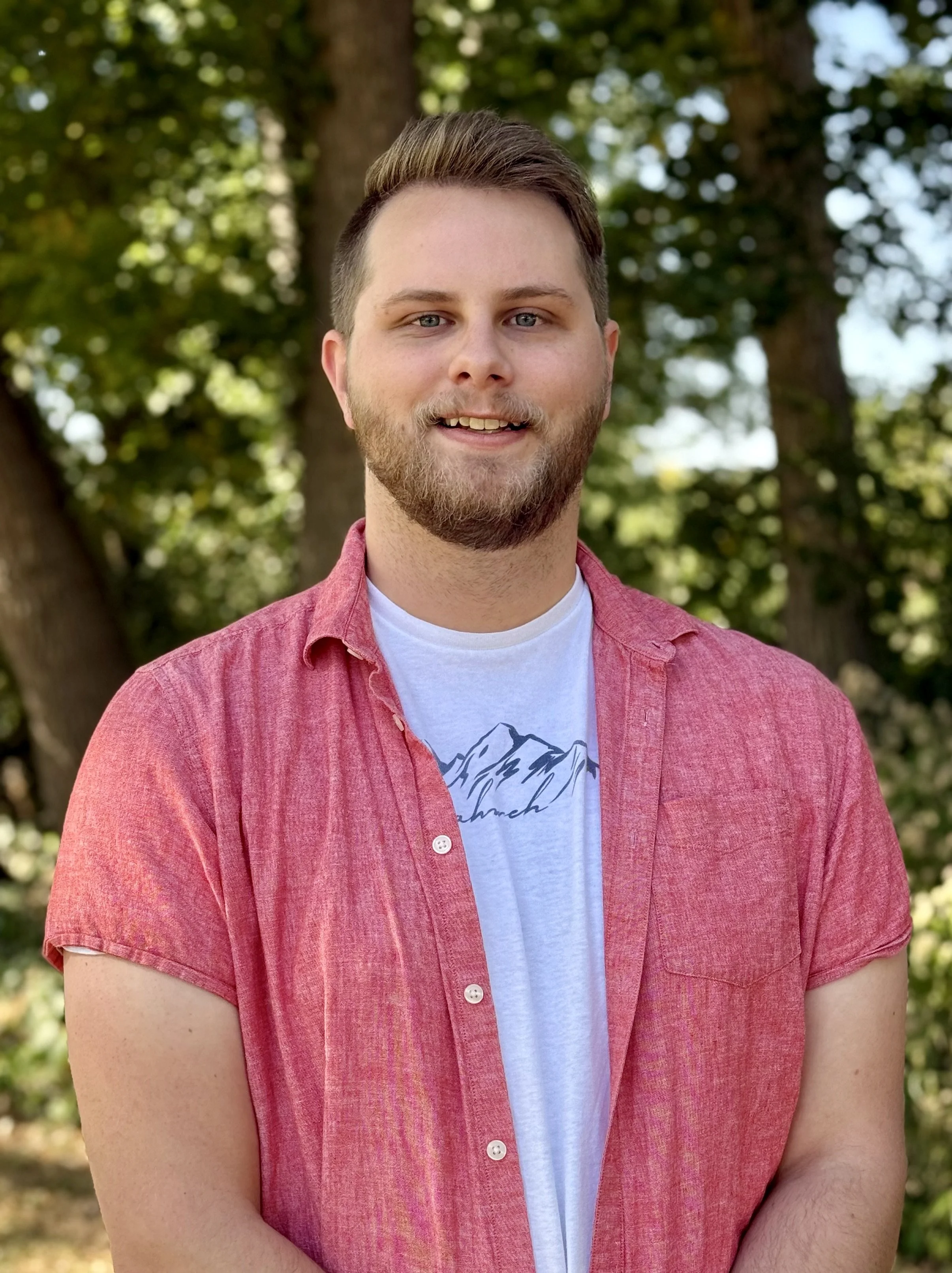 A young man with a beard and short brown hair smiling outdoors, wearing a red short-sleeved button-up shirt over a white t-shirt with a mountain design, standing in front of trees and greenery.