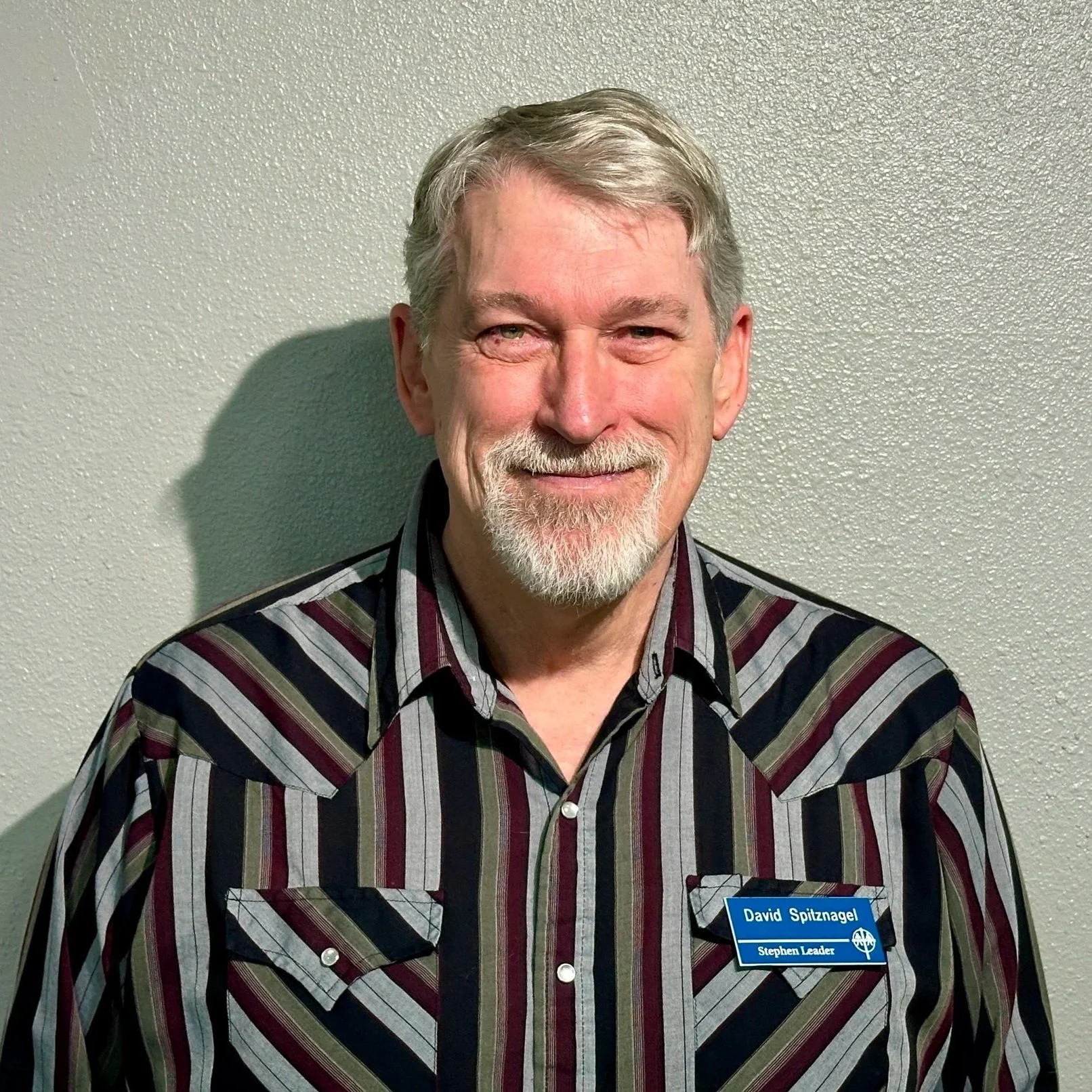 A middle-aged man with gray hair and a beard, smiling, wearing a striped shirt with a blue name tag that reads 'David Spitznagel' and 'Stephen Leader', standing against a light-colored textured wall.