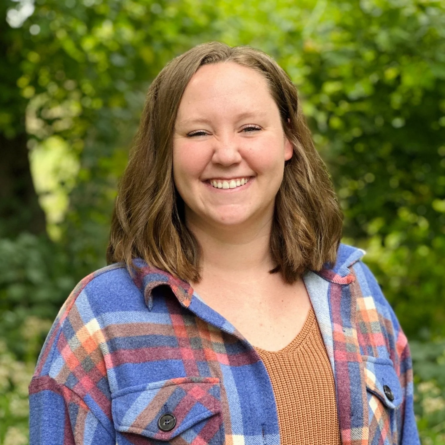 A smiling woman with shoulder-length brown hair, wearing a plaid jacket and an orange shirt, standing outdoors with green trees in the background.
