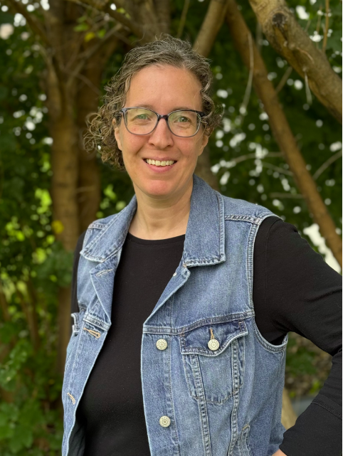 Woman with glasses and curly hair smiling outdoors, wearing a black shirt and denim vest, standing in front of trees.