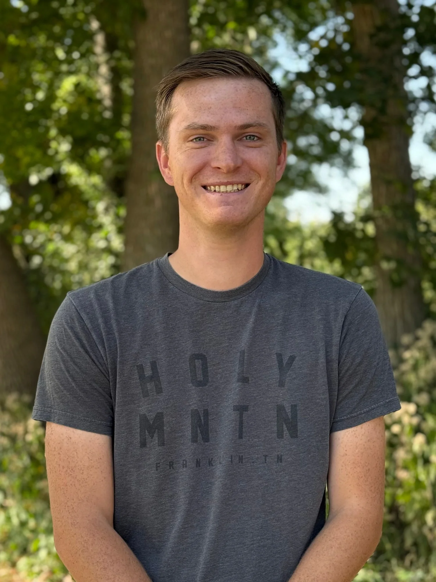 A young man with short brown hair and light skin smiling outdoors against a background of trees and greenery, wearing a dark gray t-shirt with black text.
