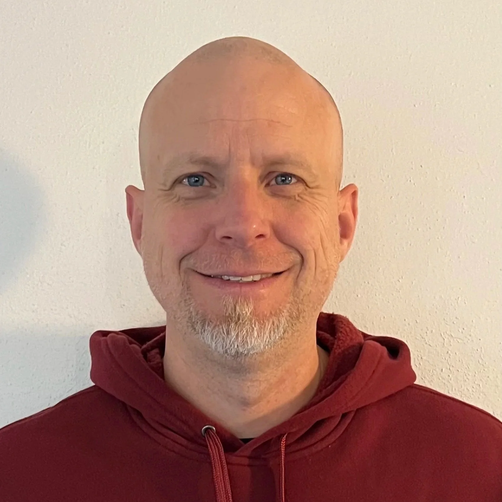 Portrait of a smiling bald man with a goatee, wearing a maroon hoodie, standing against a plain white wall.
