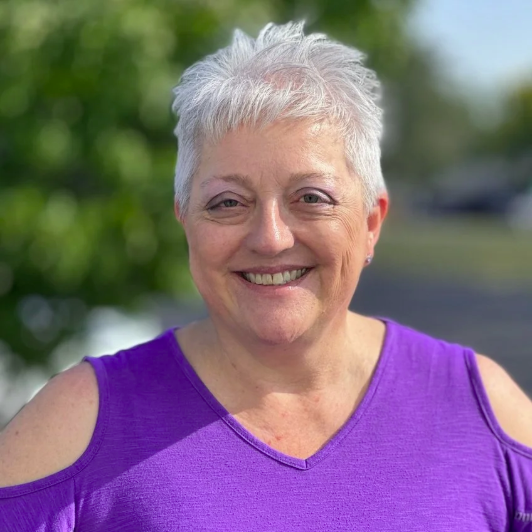 A smiling elderly woman with short white hair, wearing a purple top with cut-out shoulders, standing outdoors with blurred greenery in the background.