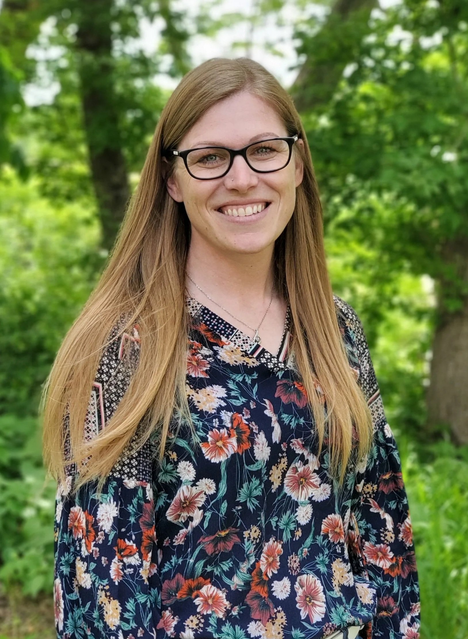 A young woman with long, straight blonde hair, wearing glasses, smiling outdoors, in front of green trees, wearing a black floral blouse and a necklace.