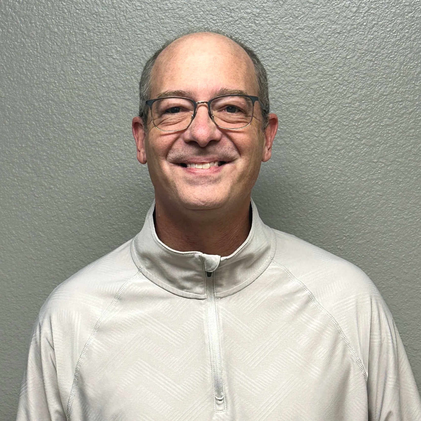 A smiling man in glasses wearing a white zip-up athletic jacket standing against a beige textured wall.