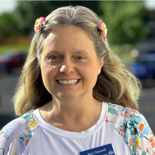 A woman with long, wavy hair wearing a floral-collared shirt and pink flower hair clips, smiling outdoors.