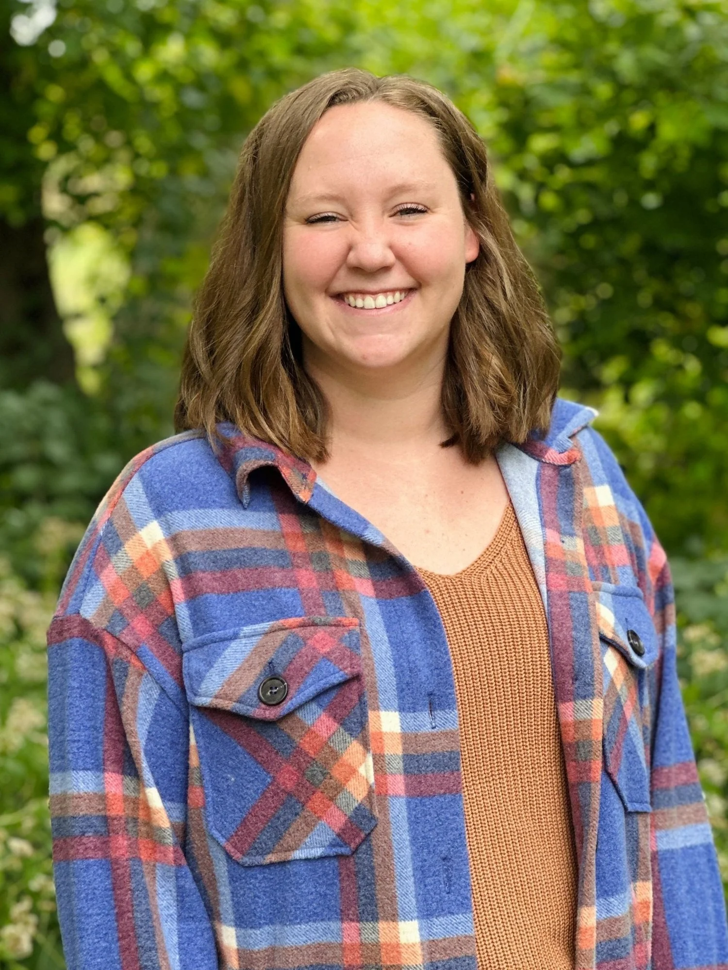 A smiling young woman with shoulder-length brown hair wearing a blue plaid jacket and a brown top, standing outdoors with green trees in the background.