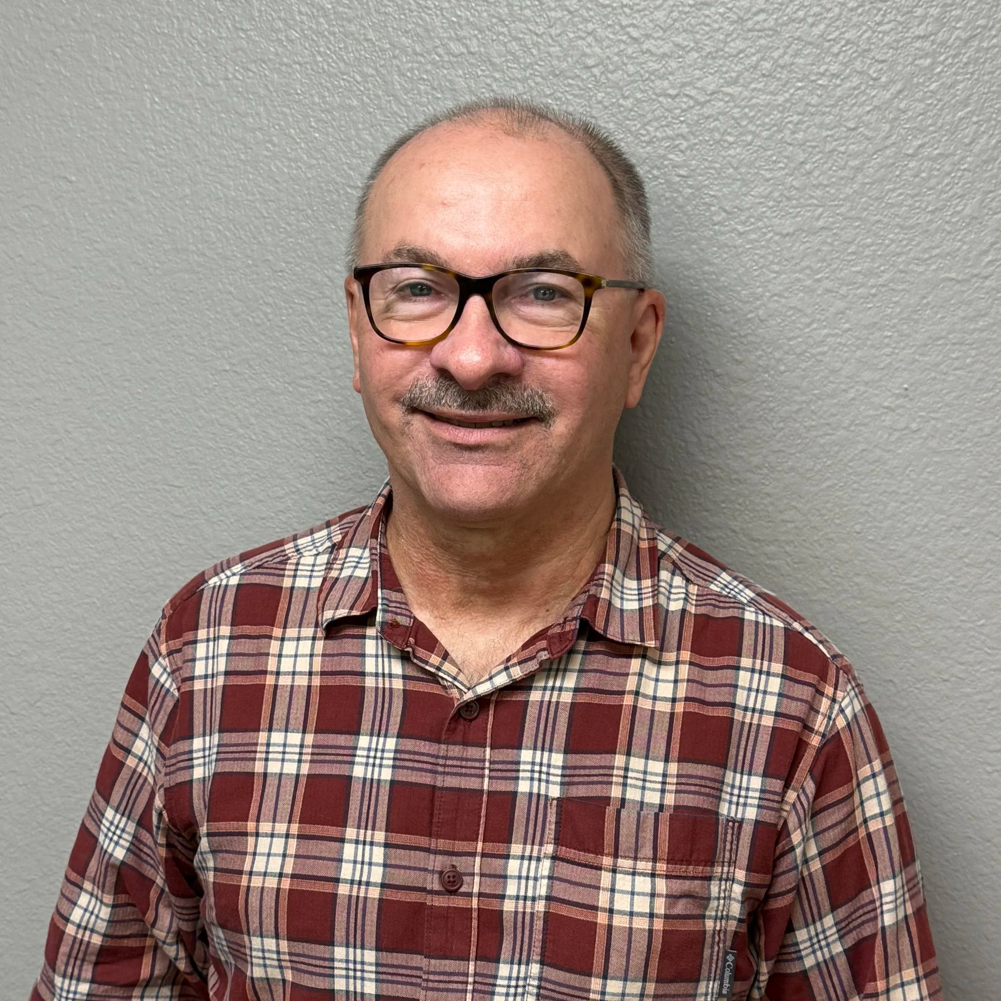 A smiling middle-aged man with glasses and a mustache, wearing a red plaid shirt, standing against a gray wall.