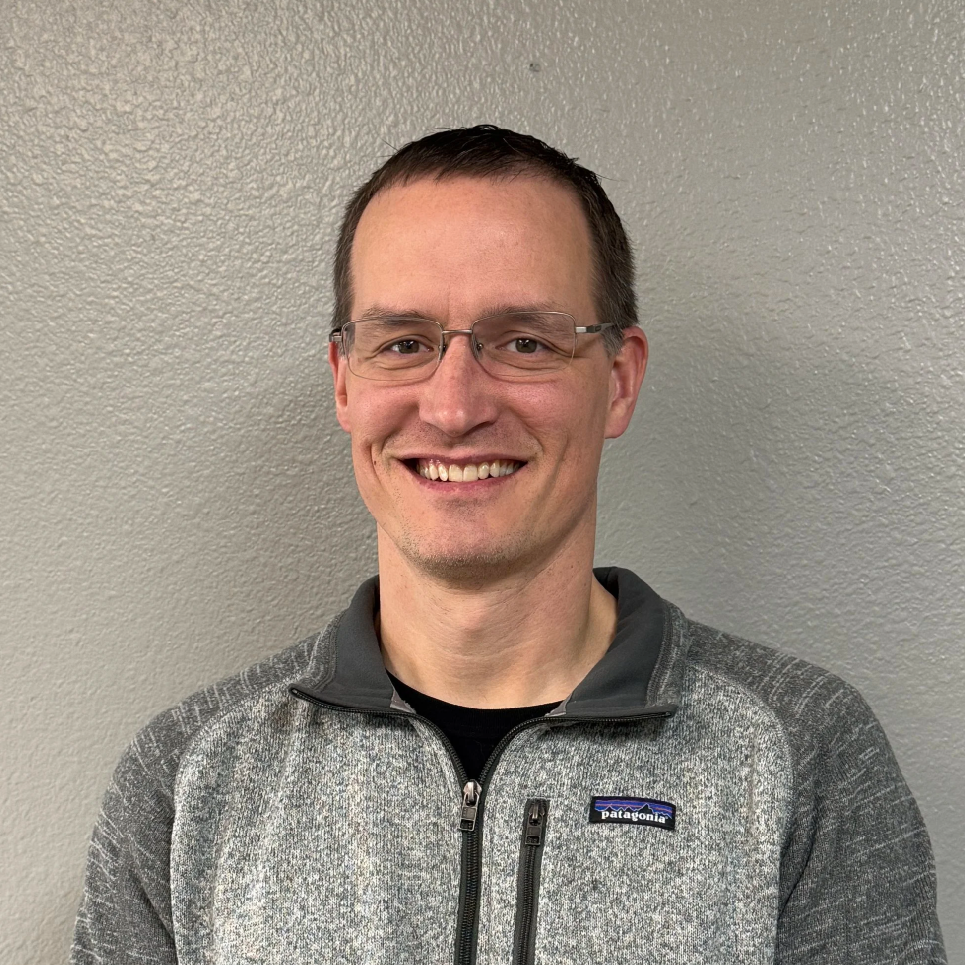 A smiling man with glasses, wearing a gray Patagonia zip-up jacket, standing against a plain gray wall.
