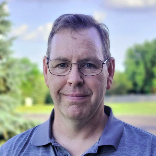 A man with short brown hair, glasses, and a slight smile, standing outdoors with trees and a blue sky in the background.
