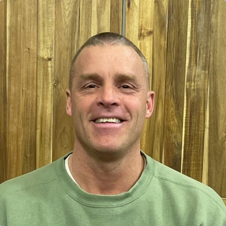 Smiling man with short hair wearing a green shirt in front of a wooden wall.