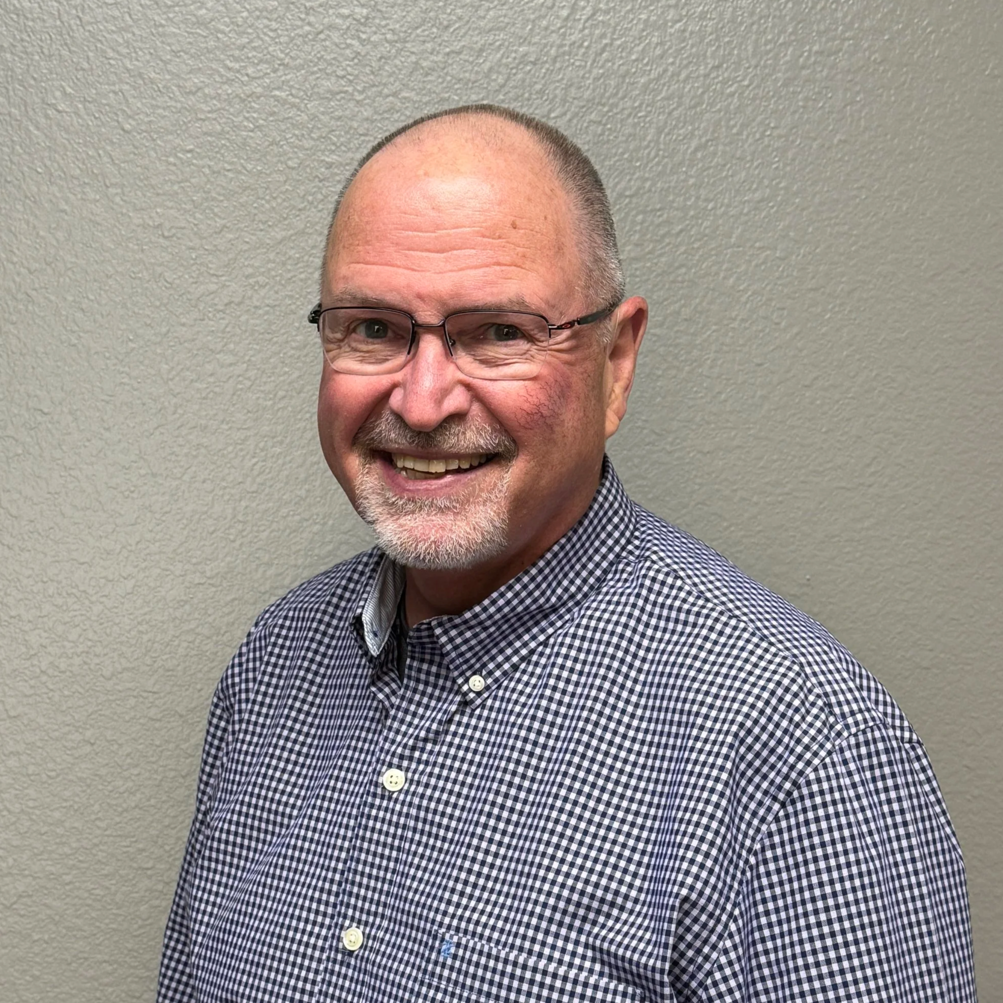 A smiling middle-aged man with glasses, a goatee, and a closely shaved head, wearing a checkered shirt, standing against a plain beige wall.