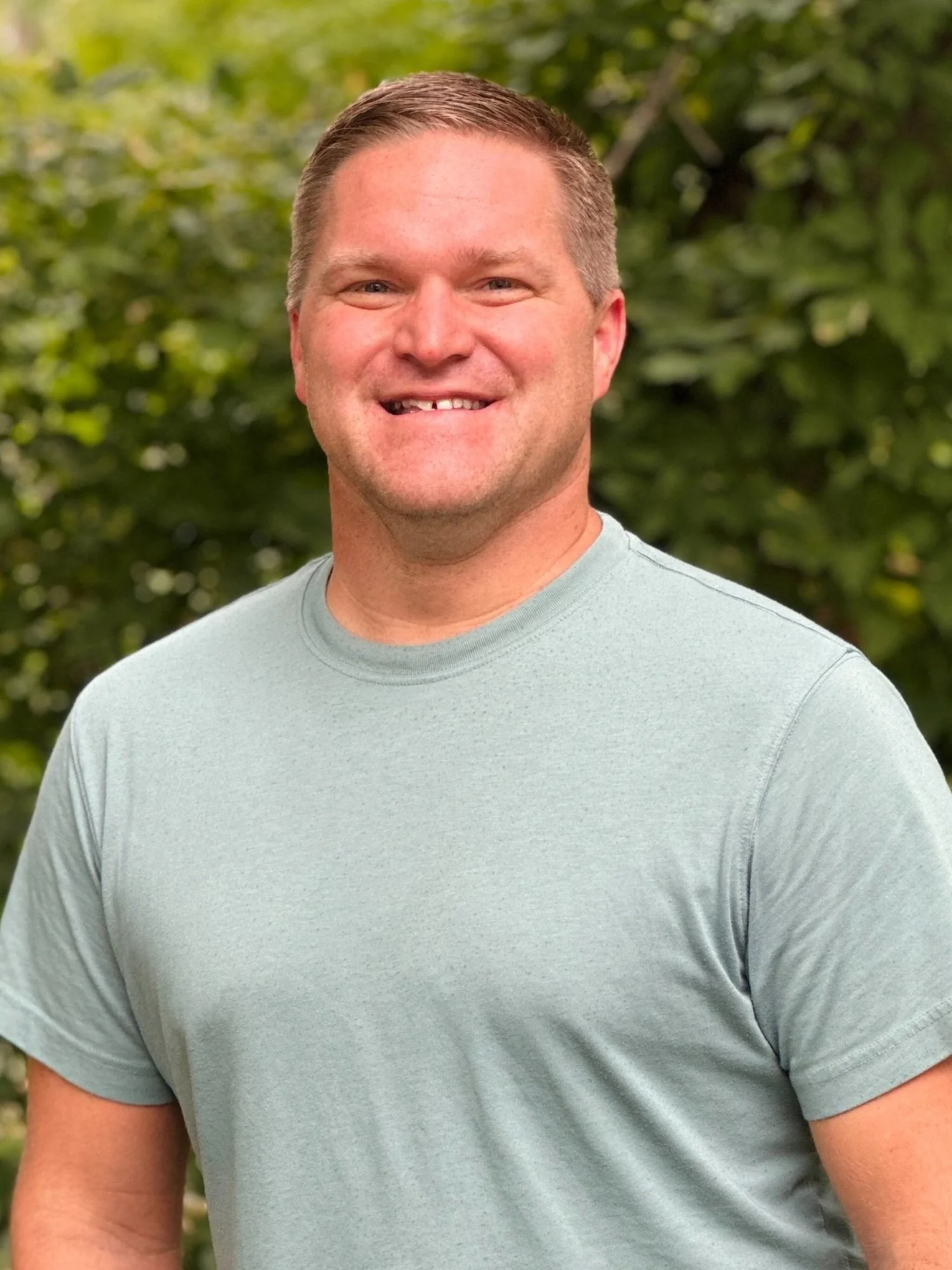 A smiling man with blonde hair and a missing front tooth, wearing a light gray T-shirt, standing outdoors with green foliage in the background.