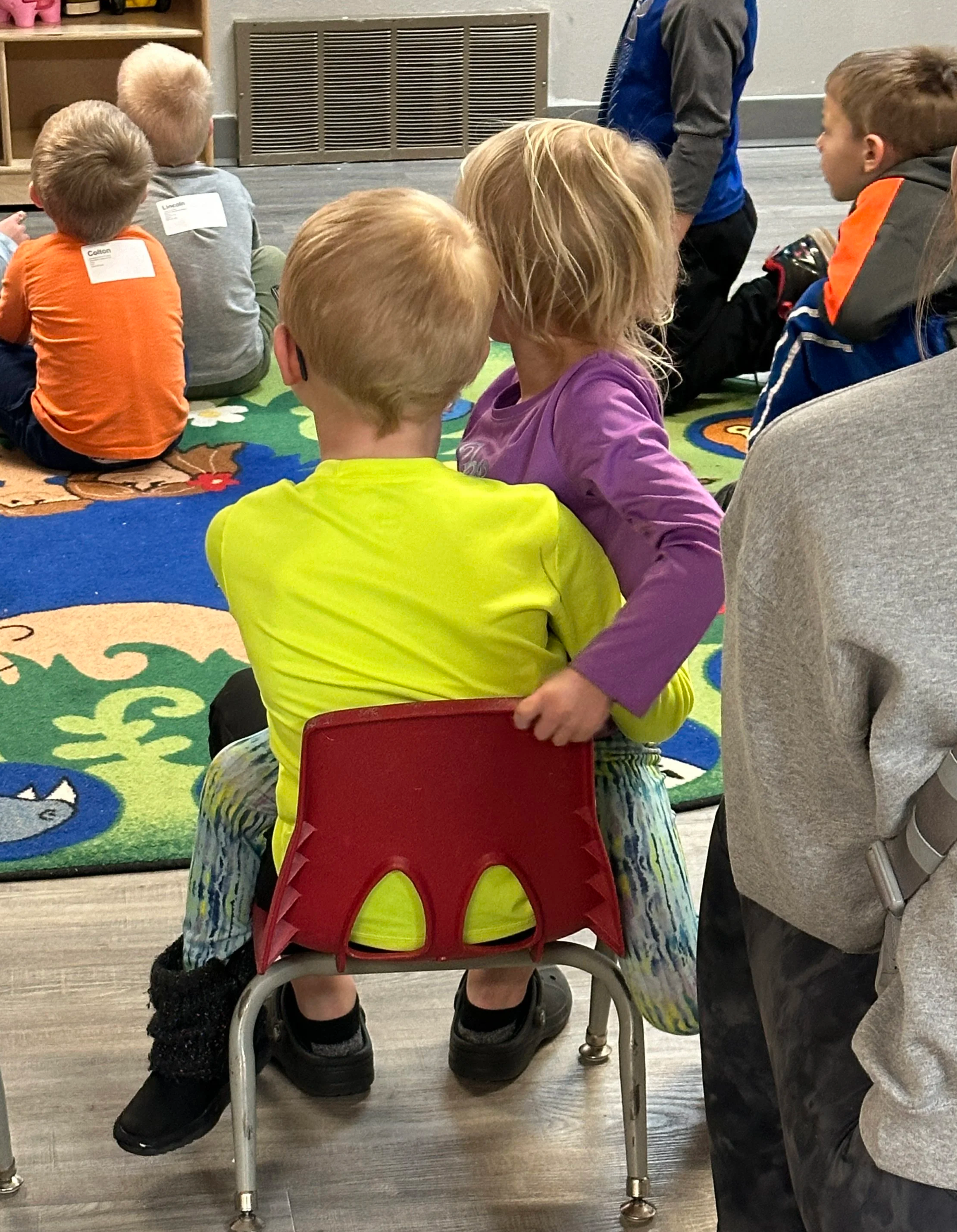 Two children sitting close together on a small red chair in a classroom with a colorful rug and other children in the background. One child, wearing a yellow shirt, is facing away from the camera, while the other child, in a purple shirt, is leaning on the first child with her arm around him.