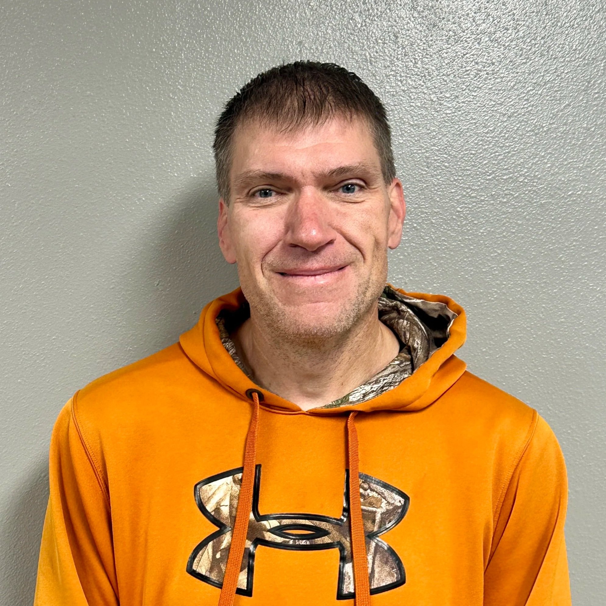 A man wearing an orange Under Armour hoodie standing against a gray wall, smiling slightly, with short brown hair and light skin.