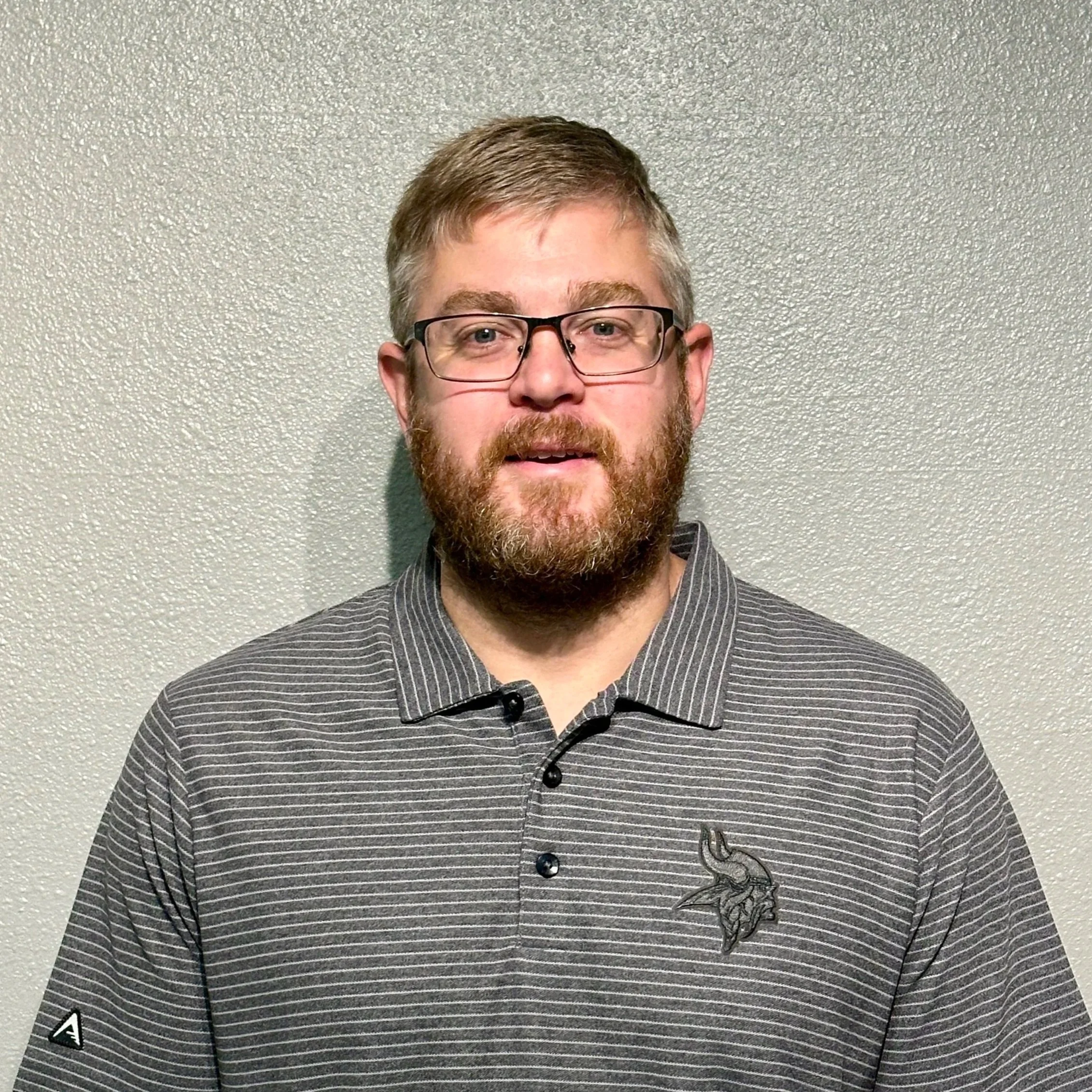 A man with glasses and a beard wearing a gray striped polo shirt with a Minnesota Vikings logo, standing in front of a gray textured wall.