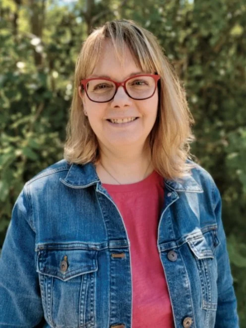 A woman with blonde hair, red glasses, wearing a denim jacket and a pink shirt outdoors with green trees in the background.