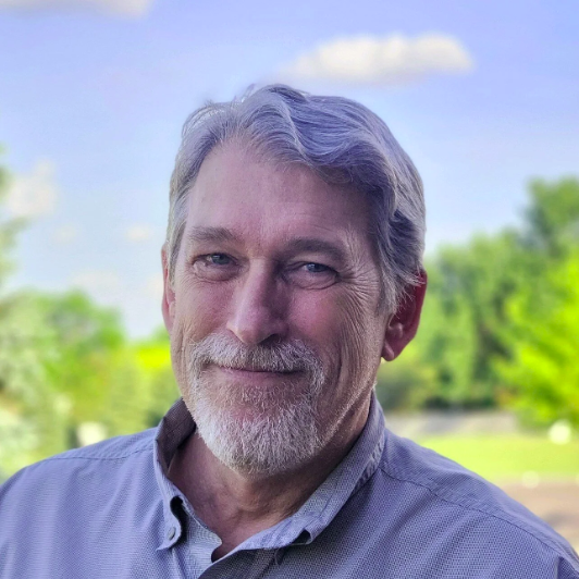 A middle-aged man with gray hair and a beard smiling outdoors with trees and a blue sky in the background.