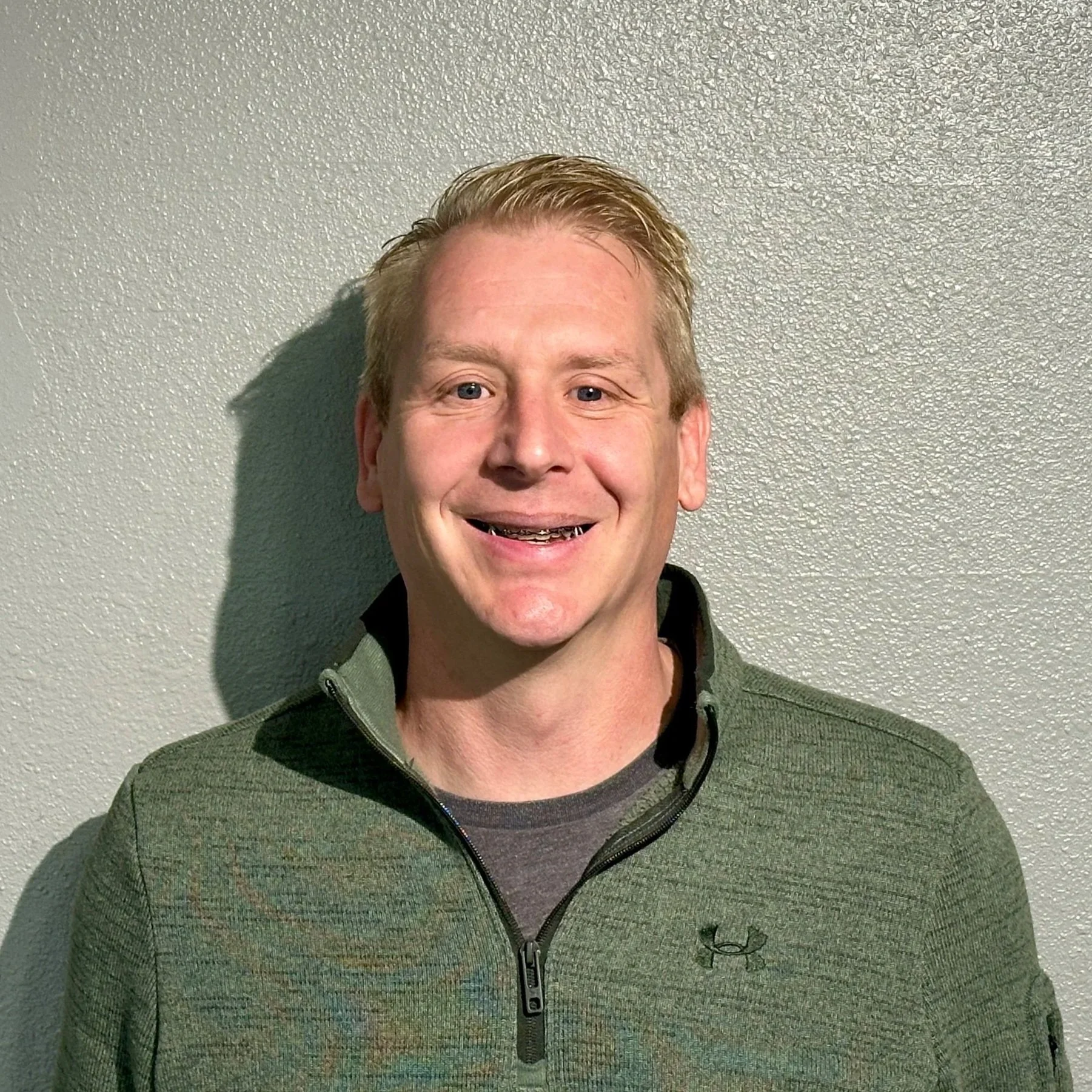 A smiling man with short blonde hair, wearing a green zip-up athletic jacket and a dark shirt underneath, standing against a textured gray wall.