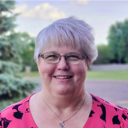 A smiling woman with short gray hair, glasses, and a pink patterned top standing outdoors with trees and sky in the background.