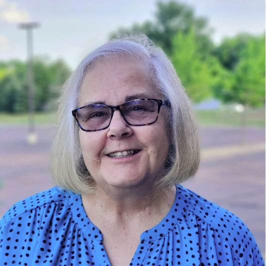 A smiling elderly woman with shoulder-length gray hair, wearing glasses and a blue polka dot blouse, standing outdoors with a blurry background of trees, sky, and a parking lot.