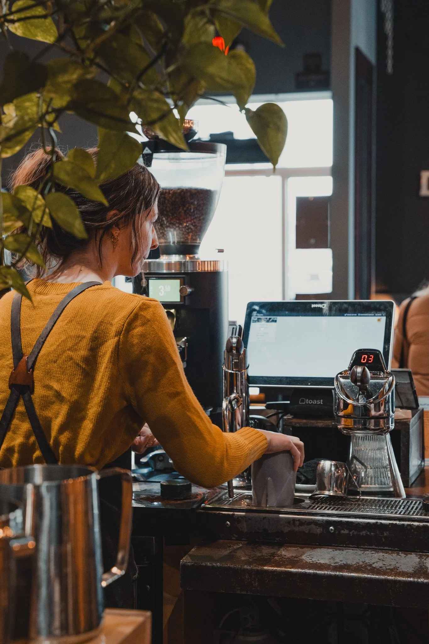 A woman in a mustard yellow sweater preparing drinks behind a coffee bar, with coffee equipment and a large glass coffee grinder visible in a cafe setting.