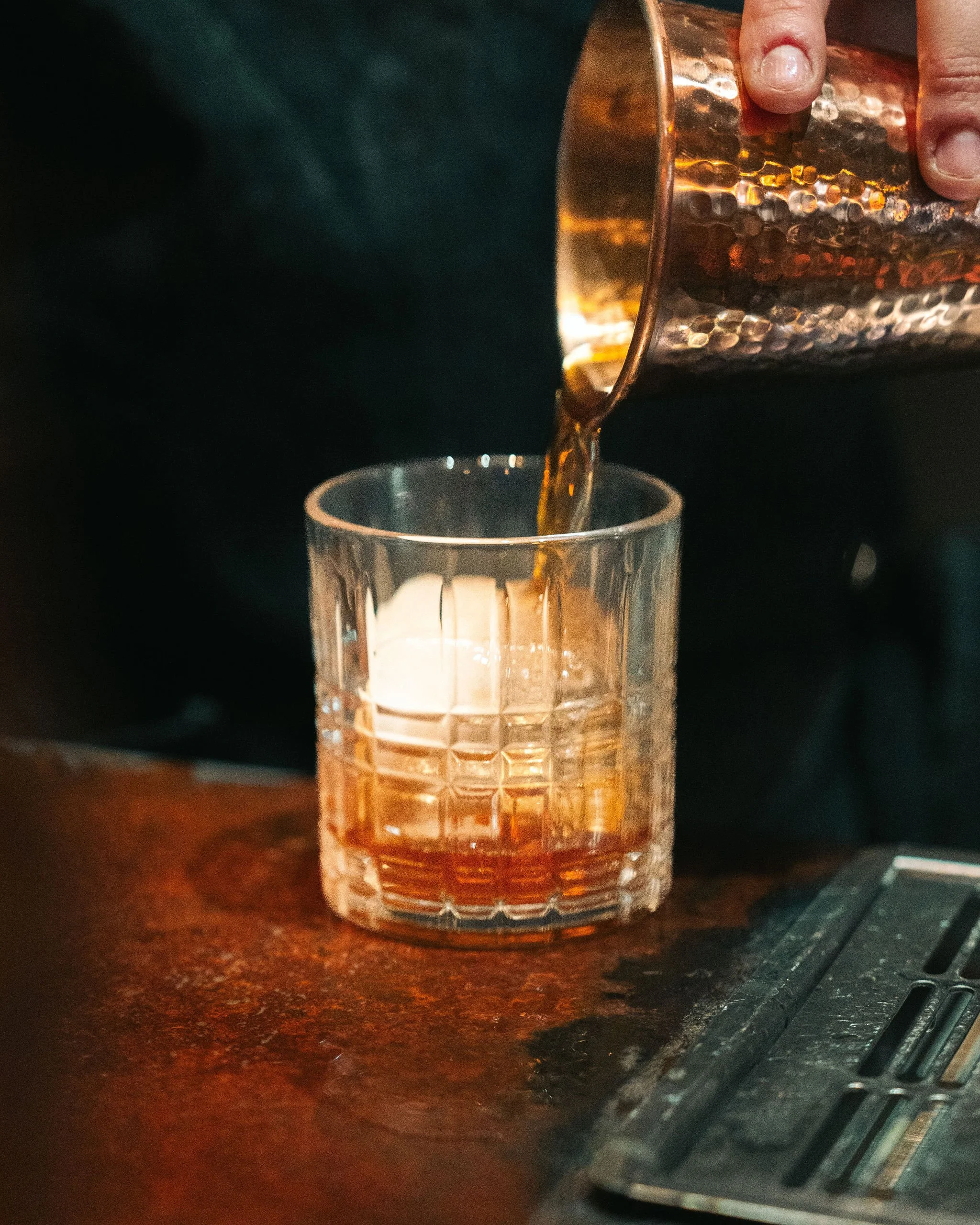 A person pouring a drink from a copper cocktail shaker into a glass on a wooden bar counter.