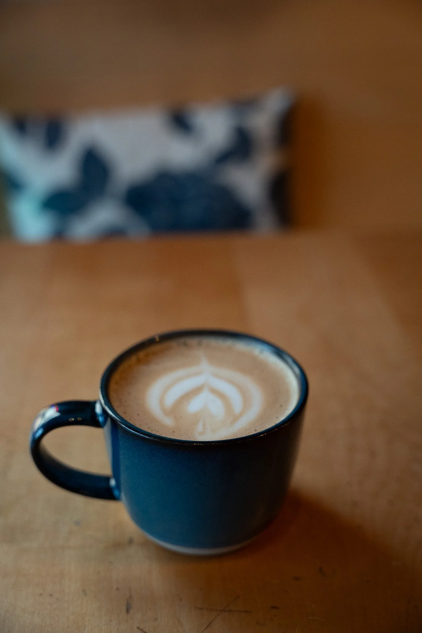 A black coffee mug with latte art on top, placed on a wooden table, with a blurred patterned pillow in the background.