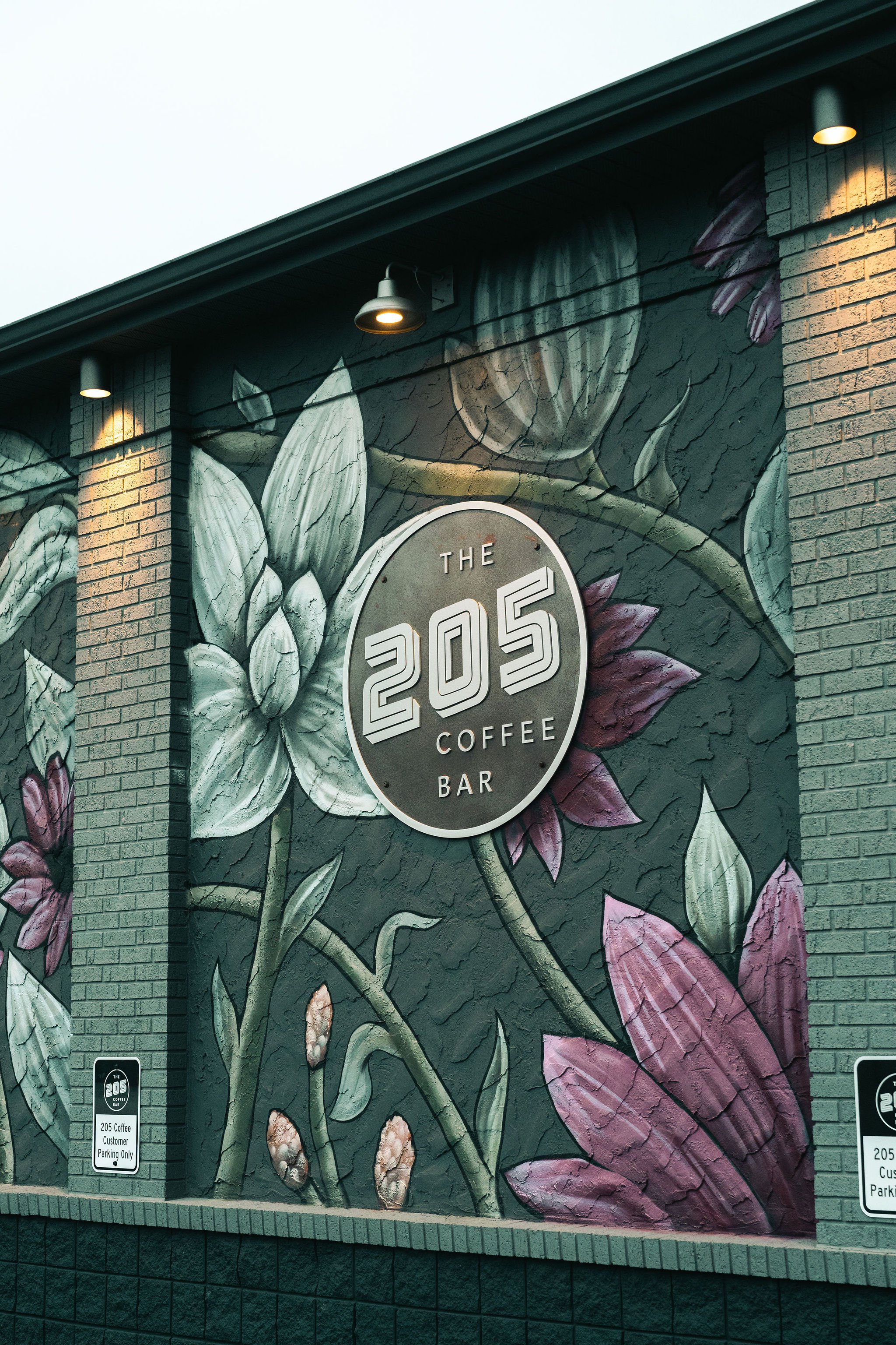 A mural on a brick wall featuring large leaves and flowers with a circular sign that reads 'The 205 Coffee Bar'.