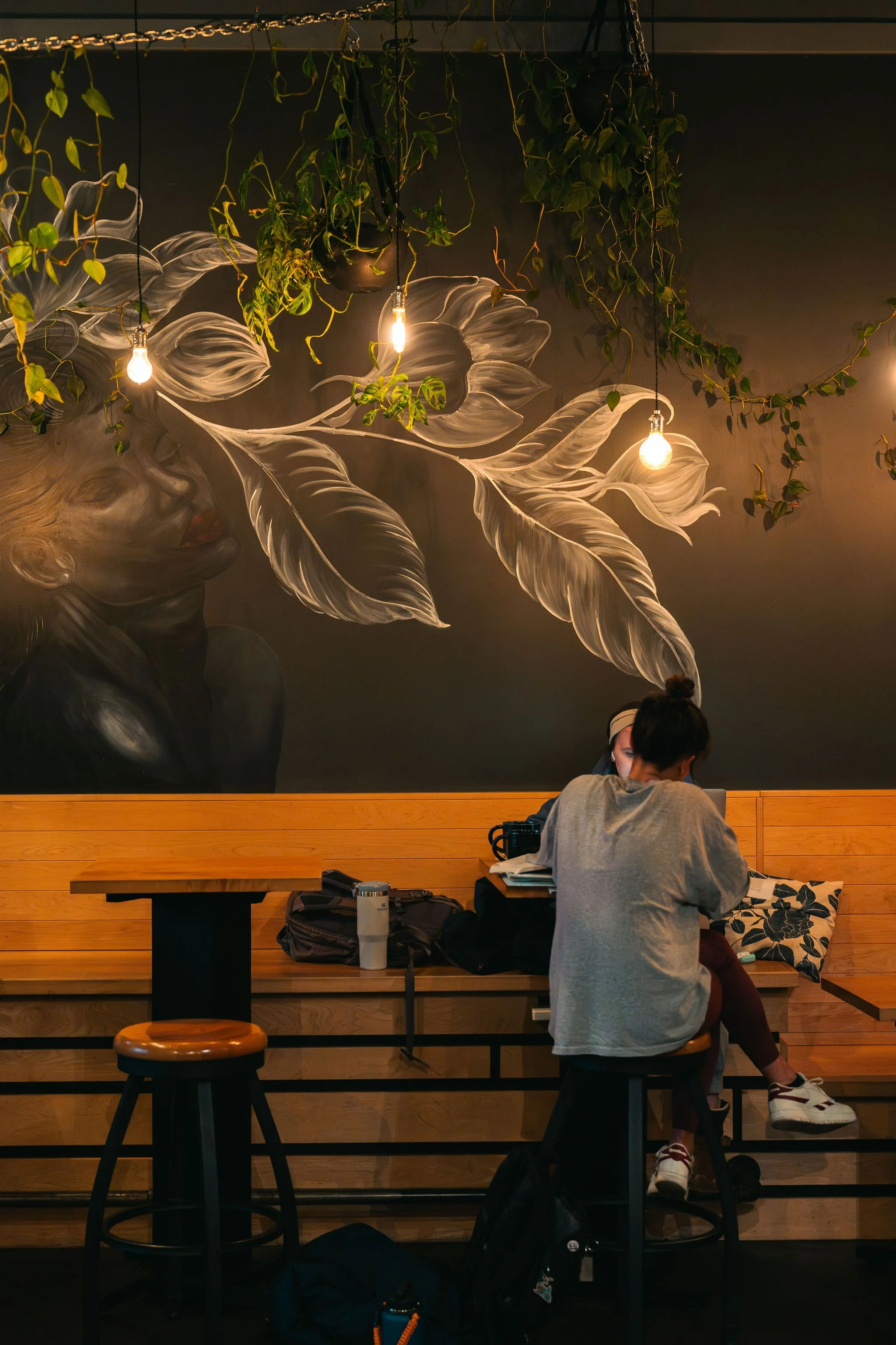 A woman sitting at a counter in a coffee shop, working on a laptop with another woman sitting across from her. The coffee shop has a mural of a woman with large leaves on the wall behind them, and hanging light bulbs and green plants decorate the spa