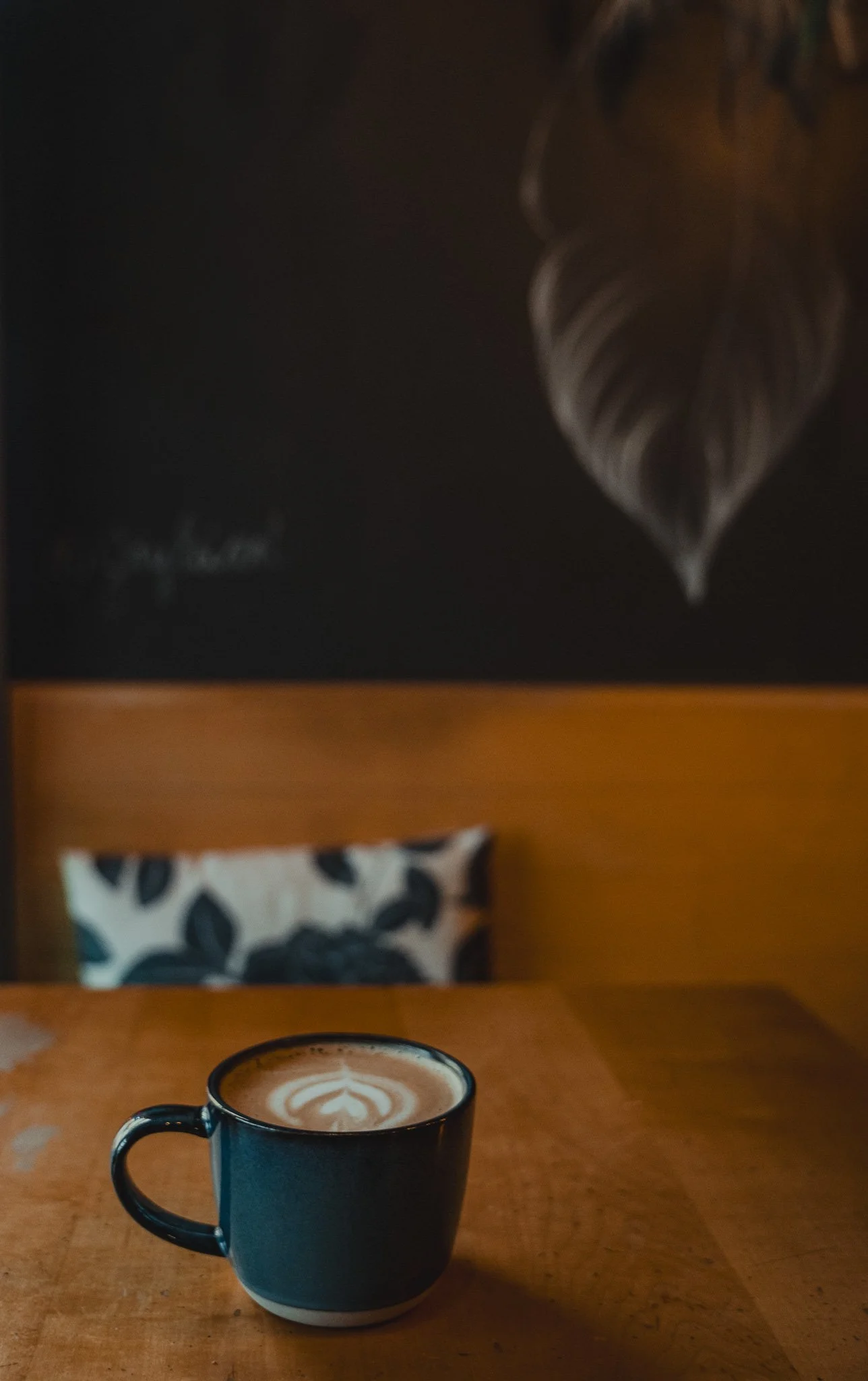 A black ceramic mug filled with a latte art coffee on a wooden table, with a blurred black and white patterned pillow and a large, detailed feather leaf artwork on a black wall in the background.