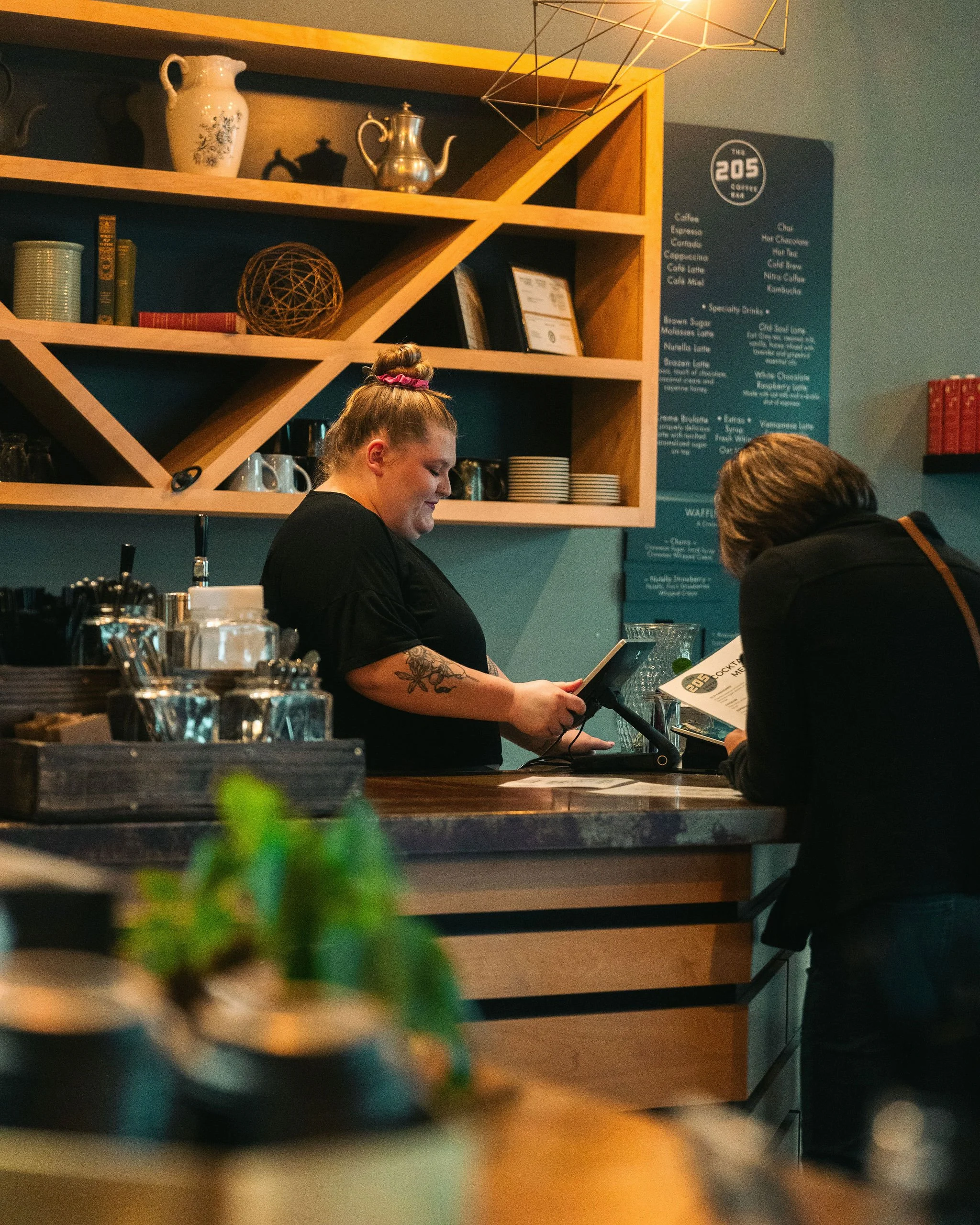 A barista and a customer at the counter of a coffee shop, with shelves of cups and a coffee menu visible in the background.