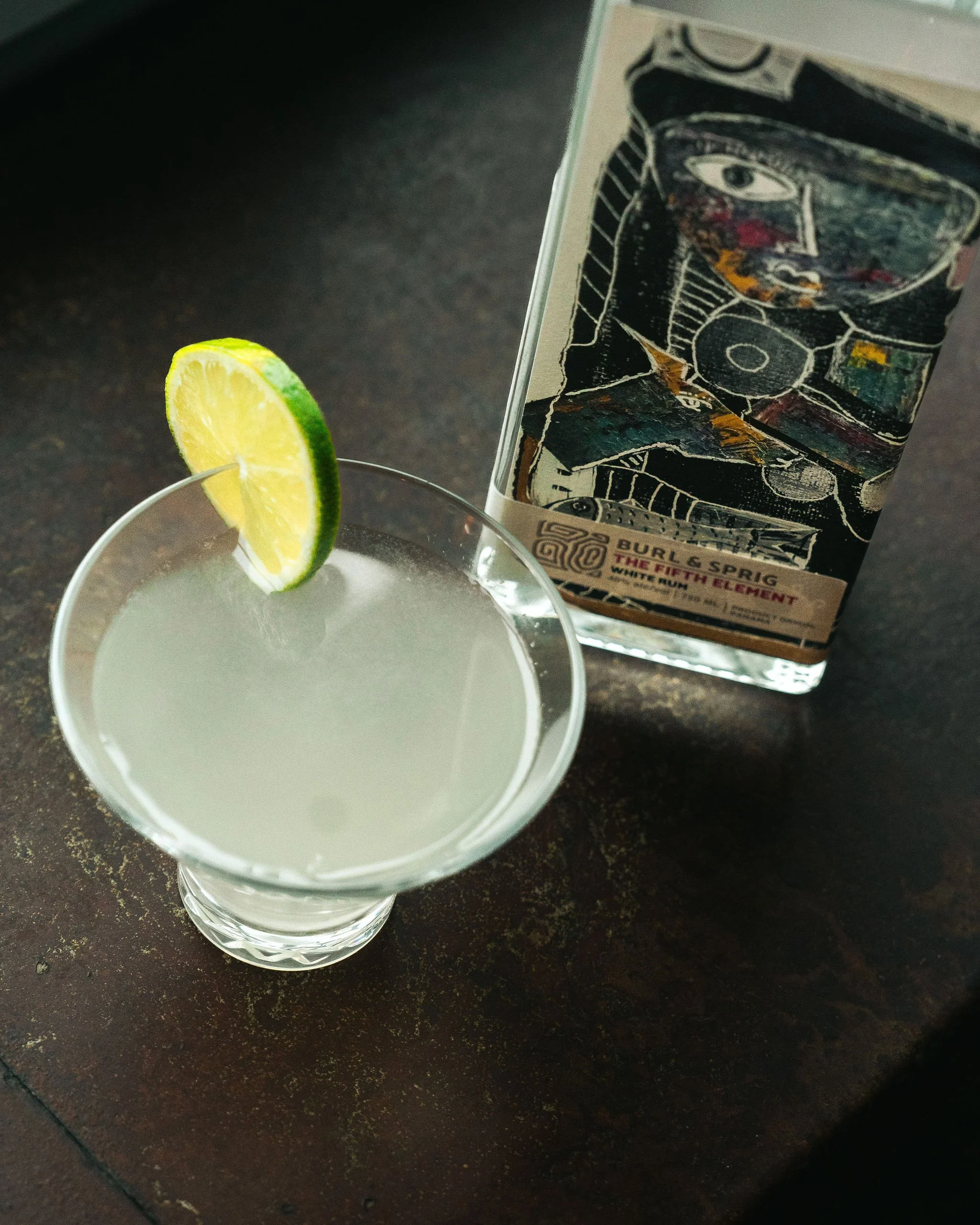A cocktail garnished with a lime wedge in a cocktail glass, with a box of Burl & Sprig Lemon Sour mix in the background on a dark surface.