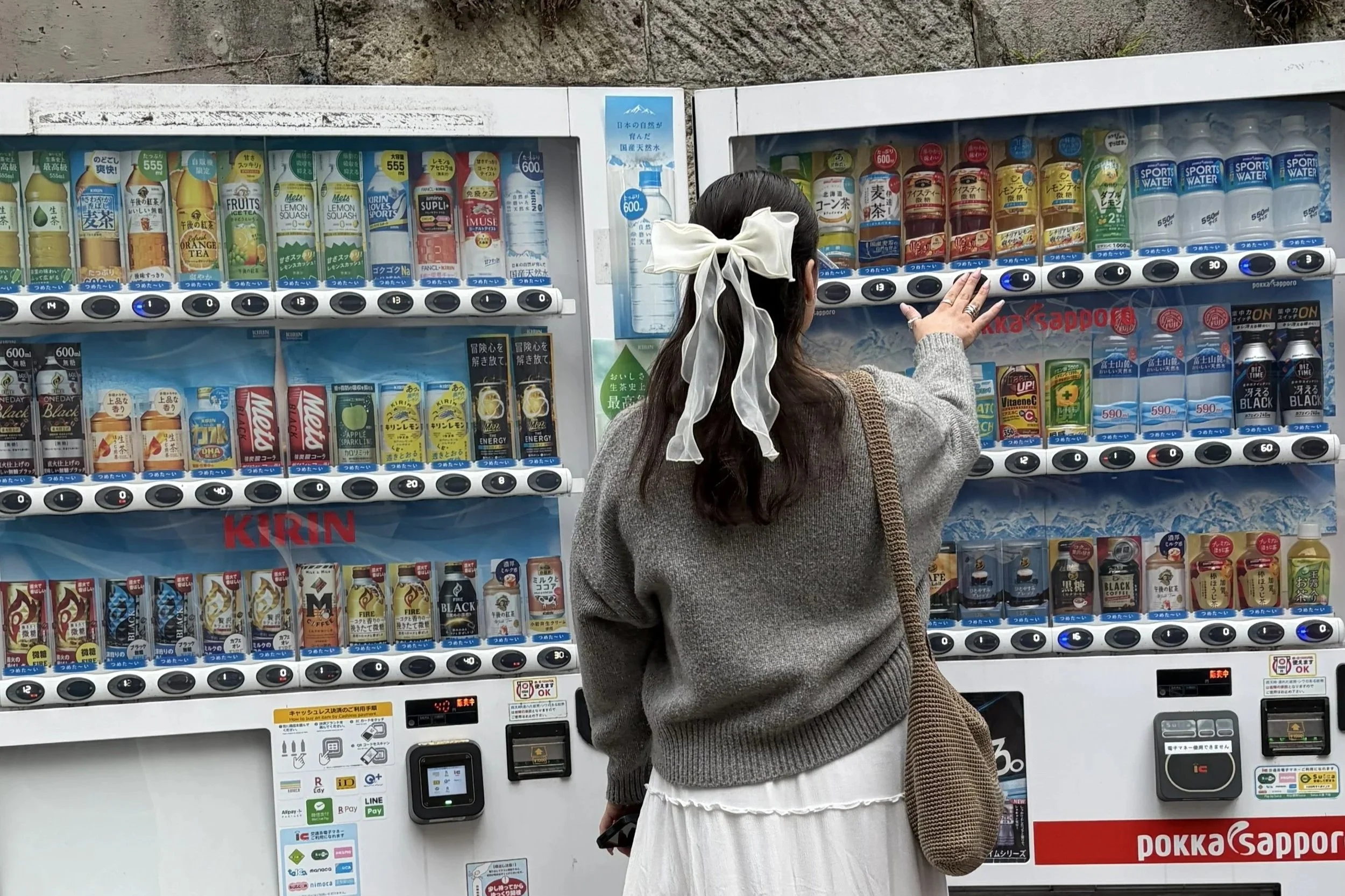A woman with dark hair, tied with a white ribbon, is reaching towards a vending machine filled with beverages, including tea, coffee, and sports drinks.