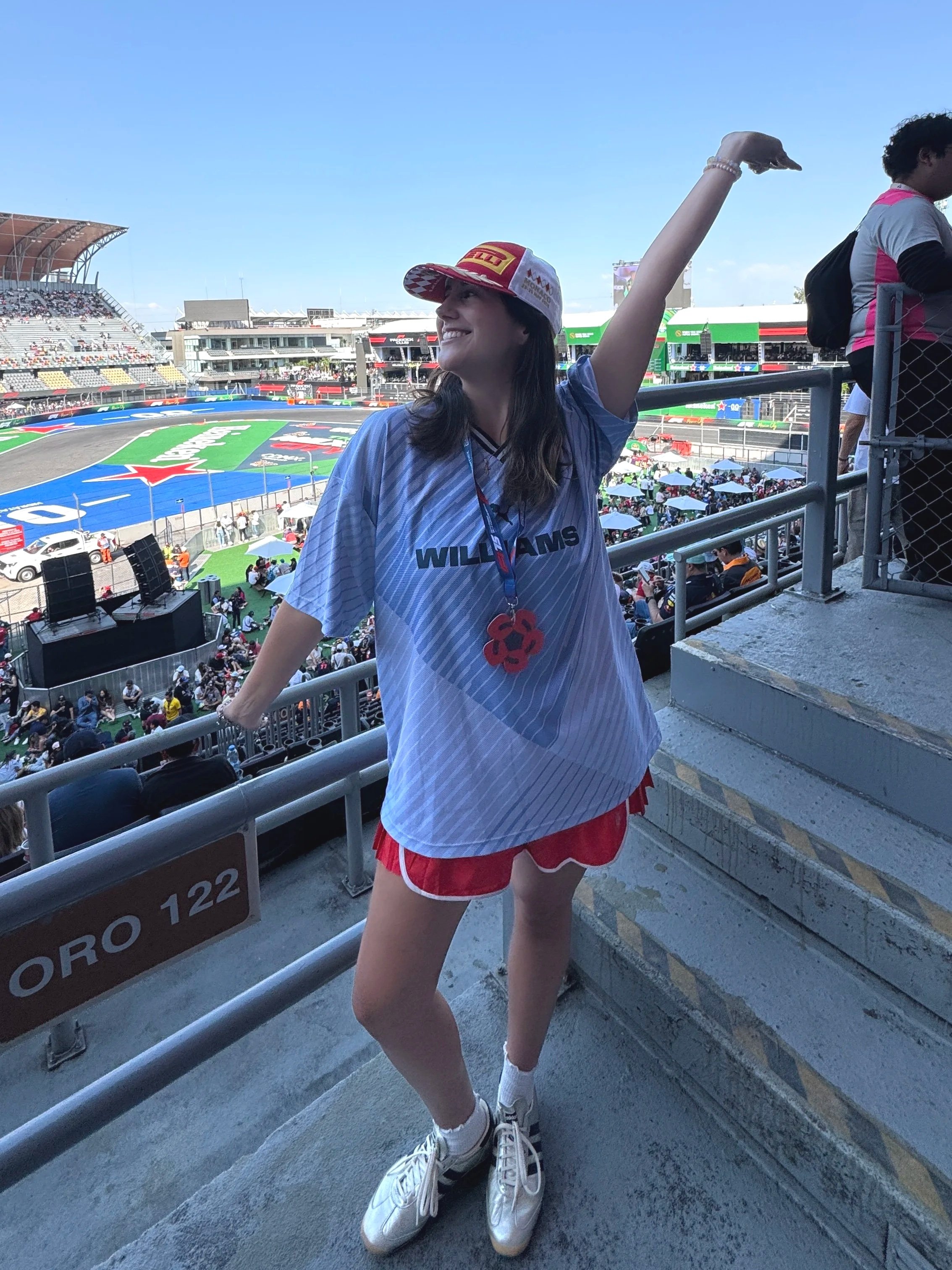 A woman wearing a Williams racing shirt, a red and white cap, white sneakers, and socks, standing at a racetrack during a sunny day with a clear blue sky, and holding her arm up.