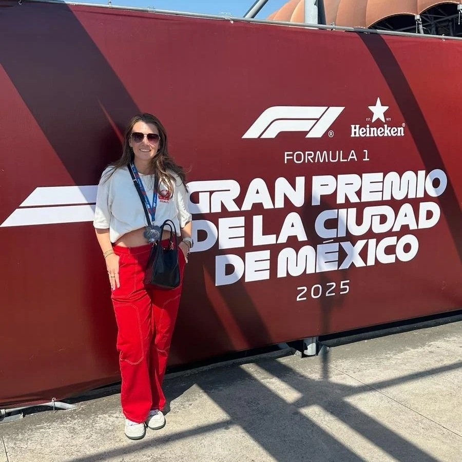 A woman wearing sunglasses, a white crop top, red pants, and white sneakers standing next to a large red banner with the Formula 1 logo, Heineken logo, and text reading "Gran Premio de la Ciudad de México 2025."