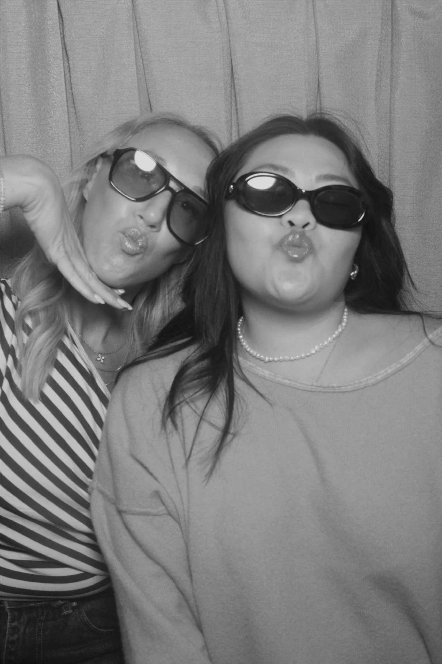 Two women wearing sunglasses, making kiss faces at the camera, with a wood-paneled wall in the background.