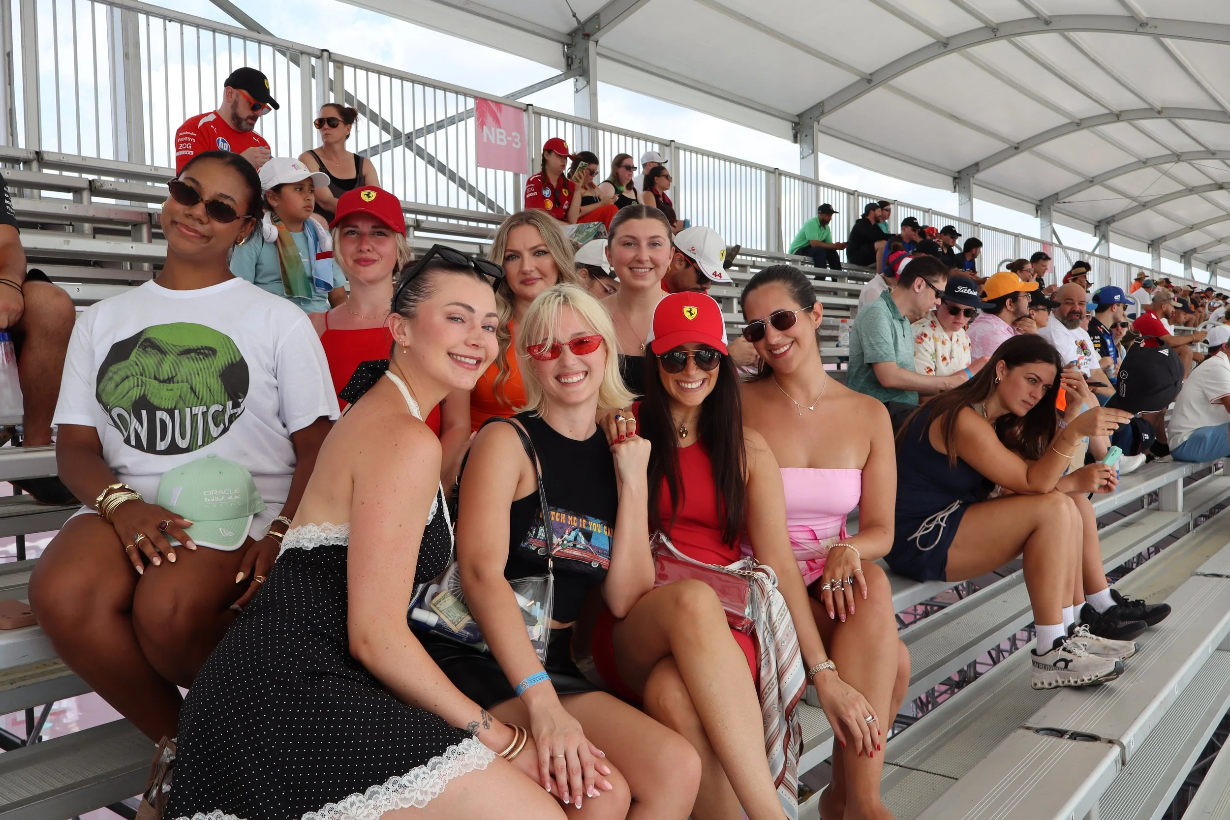 Group of people sitting on bleachers at an outdoor racing event, some wearing sunglasses, hats, and branded racing shirts, with a cloudy sky overhead.