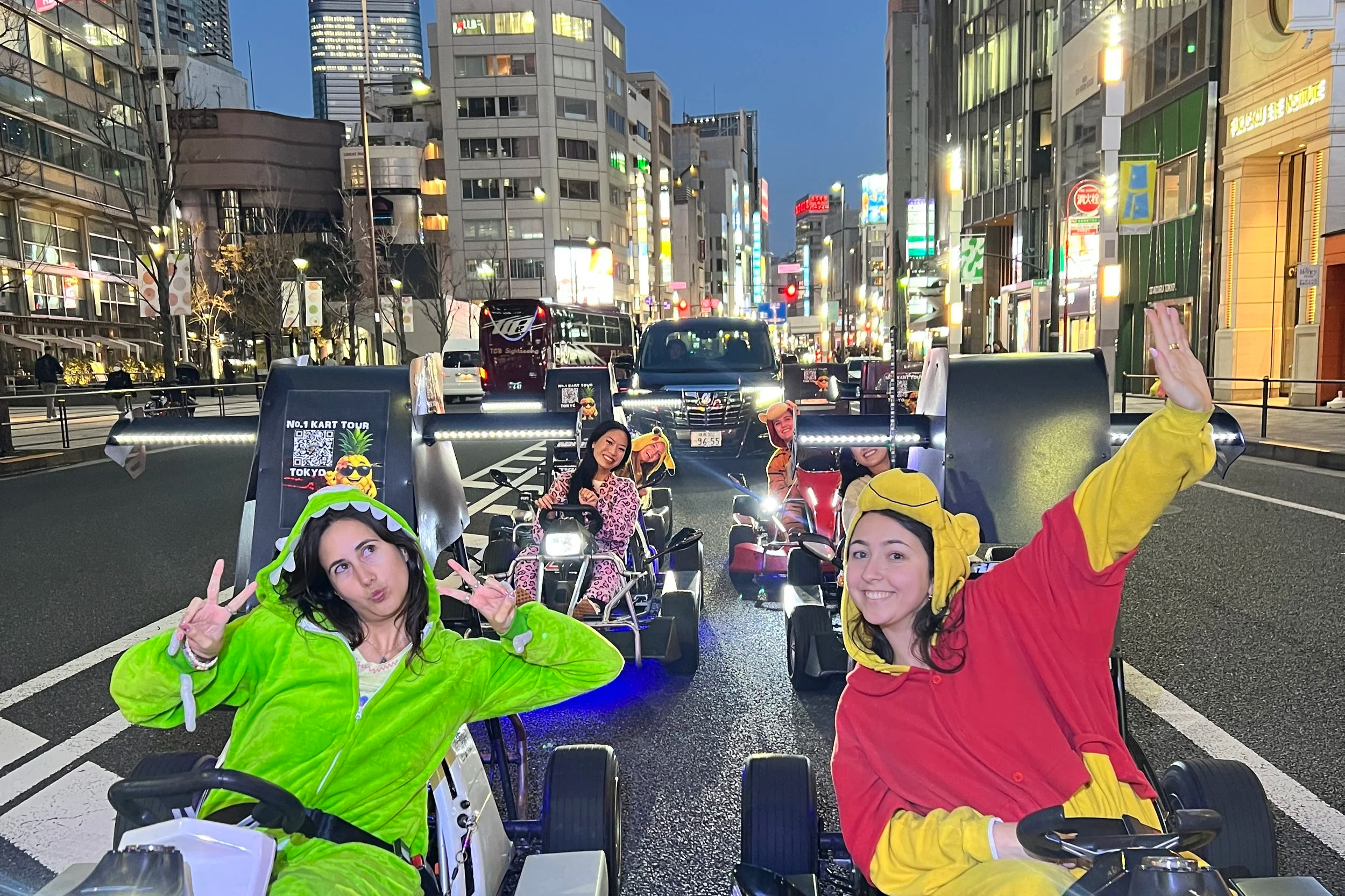 Group of young women riding electric tricycles in a city street at night, smiling and posing for the camera, with tall buildings and bright city lights in the background.