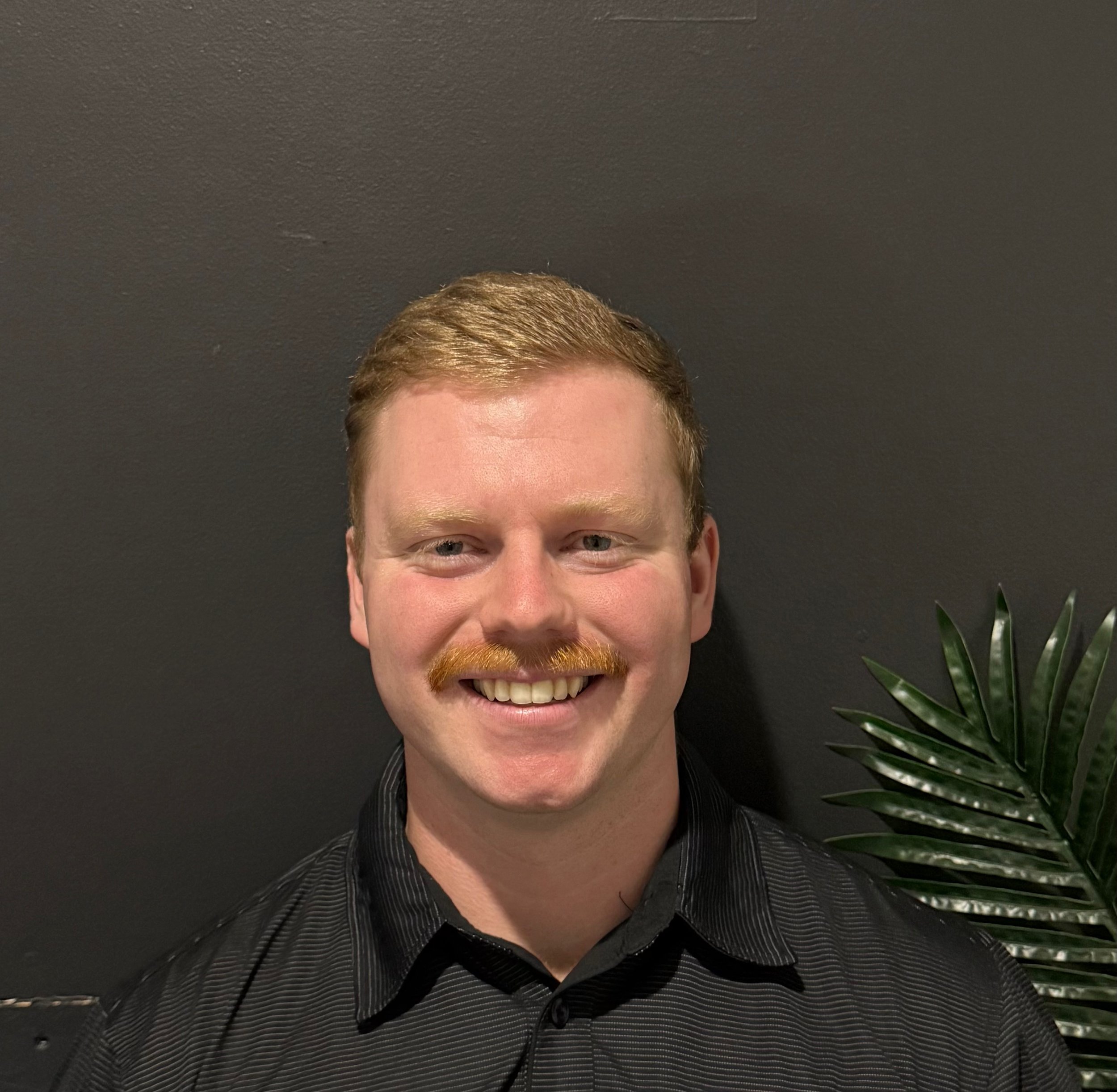 The image shows the top part of a person with light brown hair and a mustache, smiling, wearing a black and white striped shirt, with a black background and a green plant on the right side.