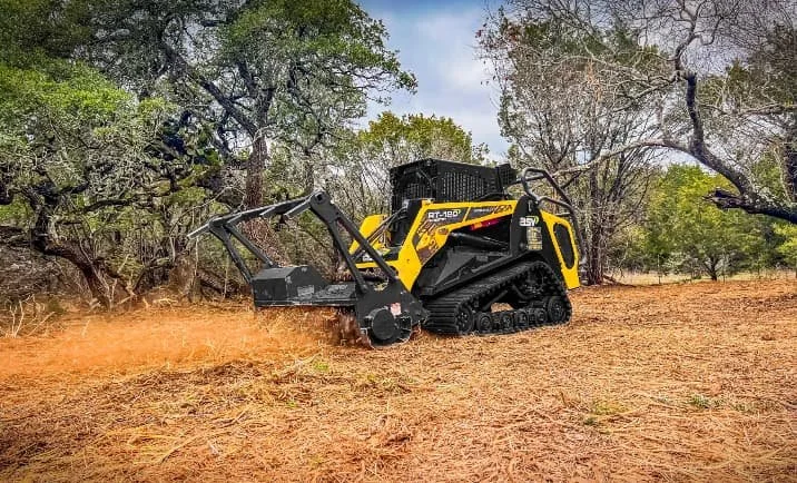 A yellow and black Bobcat compact track loader with a brush cutter attachment clearing a dry, dirt-covered area surrounded by trees.