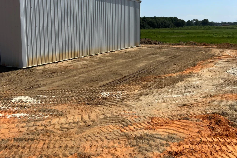 Dirt and gravel ground next to a metal building on a construction site with green fields and trees in the background.
