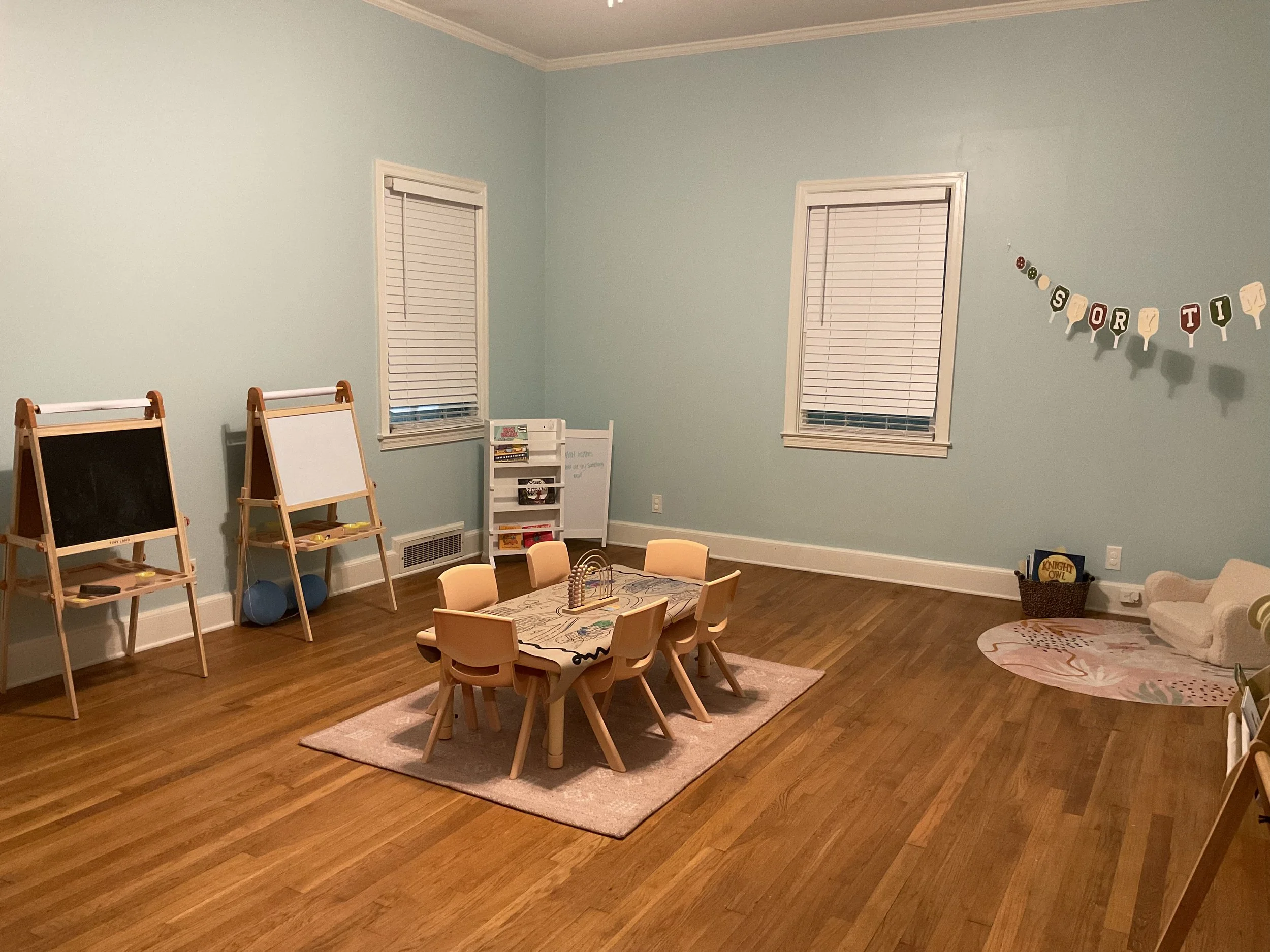 A children's playroom with two chalkboards, a small white book rack, a round pink area rug with designs, a rectangle beige rug, and a wall decoration spelling out "Storytime" with letter blocks, located near two windows with closed blinds and beige trim.