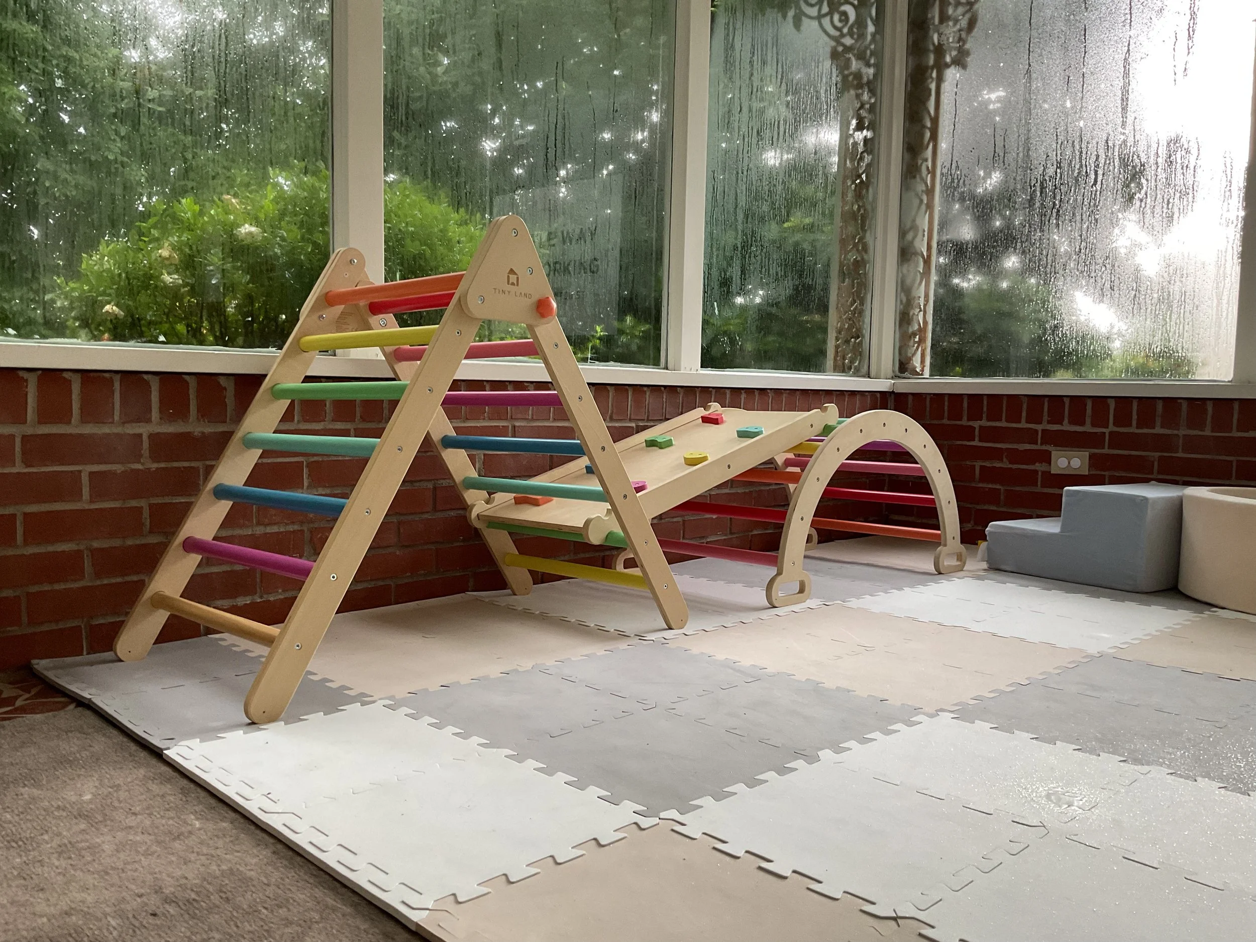 A children's indoor play area with a colorful climbing frame placed on white foam mats, located next to large rain-covered windows with an outside view of greenery and raindrops.